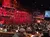 The image shows a festive event at a theater-style venue with tables, decorated with strings of colorful lights and rustic stage props, where people are gathered and dining.
