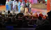 A group of performers is on stage, engaging an audience in what appears to be a theatrical production with a rustic barn-themed set.