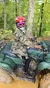 A person wearing a camouflage jacket and helmet rides an ATV in a wooded area.