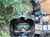 Two people wearing helmets are enjoying a ride on ATVs in a forested area, with one waving cheerfully.