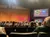 An audience is seated in a theater, facing a stage with a yellow curtain and a large screen displaying the word