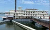 The image shows a large paddlewheel riverboat docked on a calm body of water under a partly cloudy sky.