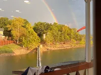 The image shows a vibrant double rainbow arcing over a river with trees on the opposite bank, viewed from a porch with decorative railing.
