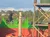 A person is chopping a log on a raised platform with a backdrop of wooden structures and colorful fencing.