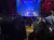 People are standing in a theater audience watching a stage with dramatic lighting and a helicopter prop.