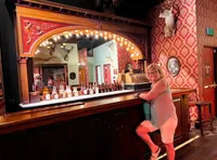 A person is standing at an ornate vintage bar adorned with bottles and surrounded by animal mounts and patterned wallpaper.
