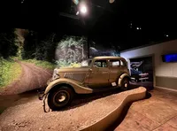 A vintage car is displayed on a gravel surface against a backdrop of a forest road in a dimly lit exhibition space.