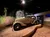 A vintage car is displayed on a gravel surface against a backdrop of a forest road in a dimly lit exhibition space.