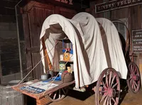 The image shows a rustic covered wagon loaded with supplies, parked in front of a wooden building labeled 