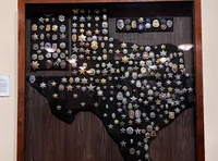 The image shows a display case containing a collection of various badges arranged in the shape of Texas, mounted on a wall.