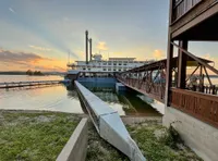 A riverboat docked at a pier is captured against a sunset backdrop, with a bridge leading from the shore to the boat.