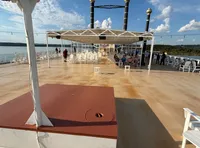 The image shows the deck of a riverboat with a covered seating area, surrounded by chairs and people, under a clear blue sky.