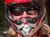 A person wearing a red helmet and mask is sitting on an ATV, with another person in the background taking a photo, next to a brick building surrounded by trees.