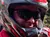 A person wearing a red helmet and sunglasses is sitting on an ATV, giving a thumbs-up gesture, with a wooded background and another ATV rider nearby.