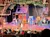 The image shows a lively stage performance with actors in colorful costumes performing in front of an audience, set against a rustic backdrop with wooden structures and trees.