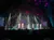 Performers in white outfits sing on a brightly lit stage with a starry backdrop, in front of an audience.