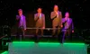 The image shows a group of performers on stage, with four men in suits singing above and three women in matching blue gowns singing below, set against a backdrop of green lighting and mist.