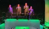 A group of three women in teal gowns sing on stage, with four men in suits performing above them, set against a green-lit, starry backdrop.