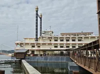 The image shows a large paddle steamer boat docked at a pier under a cloudy sky.