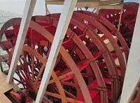 The image shows a red paddlewheel on a boat with a scenic background of a lake and distant hills.
