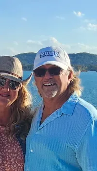 A couple is standing on a boat deck on a sunny day, enjoying the scenic view of the water and distant hills.