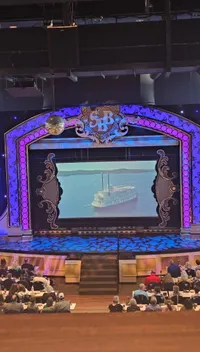 The image shows an ornate theater stage with a large screen displaying a steamboat, surrounded by an audience seated at tables.