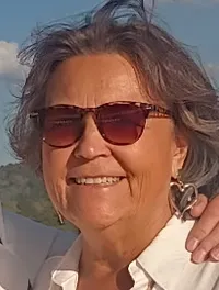 A couple stands smiling and embracing on a sunny deck, with a scenic view of water and trees in the background.
