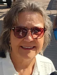 A man and a woman wearing sunglasses are taking a selfie on a sunny day with a waterfront and structures in the background.