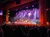 A lively stage performance is taking place with colorful lighting, a live band, and a seated audience, under the banner 