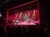 A live performance on stage features dancers and musicians under dramatic red and white lighting.