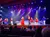 A group of performers in colorful, glittery costumes are singing and dancing on a brightly lit stage in front of an audience.