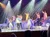A group of performers sing and dance on a stage with colorful lighting effects while an audience watches.