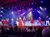 Performers in colorful costumes sing and dance on stage under bright lights, entertaining an audience.