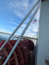 The image shows a red paddlewheel of a riverboat with an American flag flying above, set against a backdrop of water and a blue sky with some clouds.