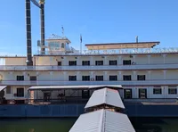 A large, multi-level riverboat with ornate detailing is docked by a covered walkway under a clear blue sky.
