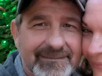 A couple is smiling in front of colorful, decorated Christmas trees and a 