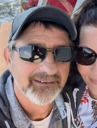 A couple wearing sunglasses is smiling for a selfie on a boat with a red paddlewheel and water in the background.