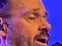 A group of six men in sparkly jackets are performing on a stage with bright lights and smoke effects.