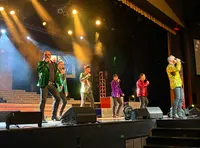 A group of performers in colorful, sequined jackets are singing on stage under bright lights.