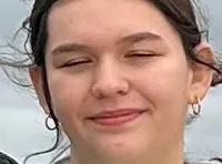 A group of three people is standing on a waterfront deck, with an American flag and a viewing telescope nearby.