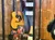 The image shows a display case containing a colorful shirt and vest, a guitar, photos, and other memorabilia against a wooden wall background.