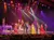 A festive stage performance featuring musicians and singers in colorful, elegant outfits under vibrant lighting and holiday decorations.