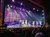 A lively stage performance is taking place at the Grand Ole Opry, featuring singers and dancers in colorful costumes, under bright stage lights.