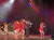 Performers in red outfits dance energetically on a stage, accompanied by a musician playing the violin.