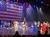 A group of performers in patriotic attire sing and dance on stage with an American flag backdrop and a live band accompanying them.