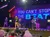 Performers are on stage in front of an audience with colorful lights and a large screen displaying the phrase 