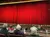 People are seated in a theater facing a large stage with closed red curtains and warm lighting.