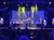A live performance is taking place on stage with singers and musicians under colorful lights, alongside a large Elvis Presley themed backdrop.