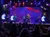A band is performing on a brightly lit stage with colorful lights and video screens, entertaining a seated audience.