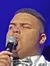 A person in a shiny white jacket and bow tie is singing into a microphone on stage.
